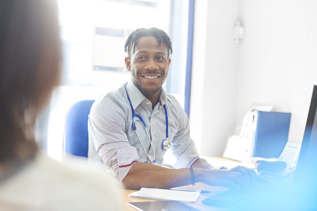 A gp wearing a stethoscope around his neck, talks to a patient who is shown with back towards camera in a blurred image