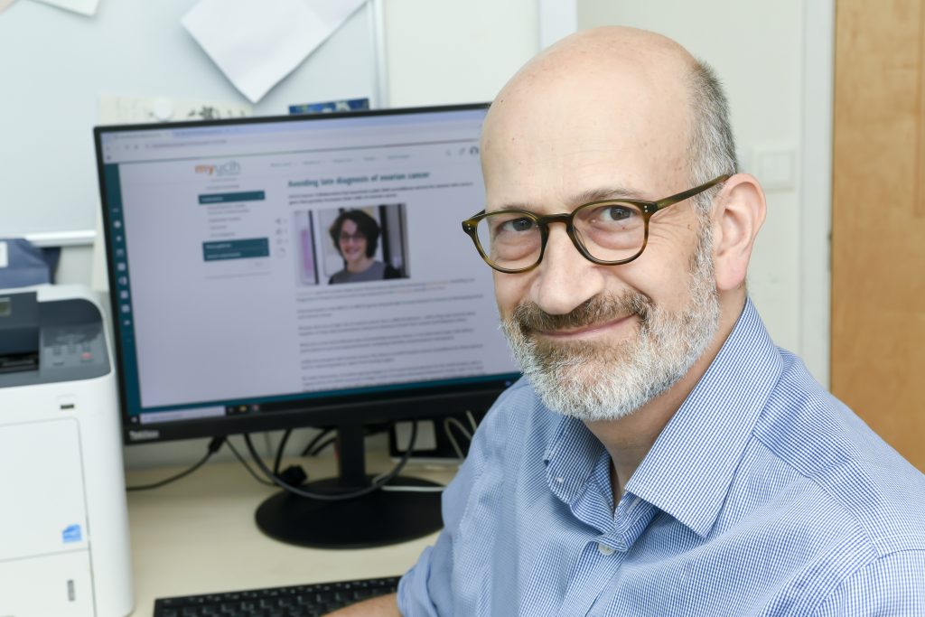 Professor Adam Rosenthal is at a desk, facing the camera, with a computer screen behind him. On the screen in blurred text the headline says Avoiding Late Diagnosis of Ovarian Cancer