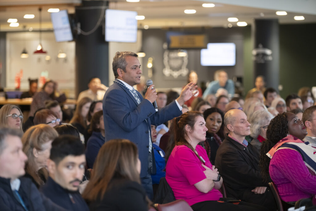 A male conference attendee, holding a microphone, stands up from the audience to speak