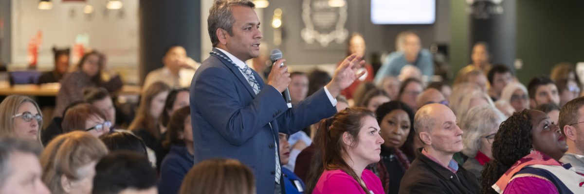 A male conference attendee, holding a microphone, stands up from the audience to speak